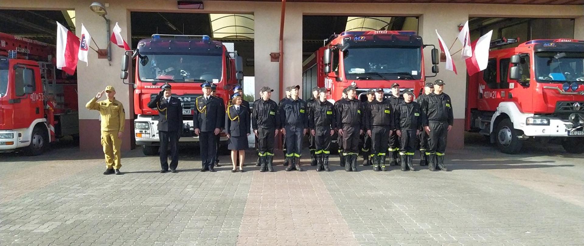 Zdjęcie przedstawia funkcjonariuszy KP i JRG podczas uroczystości wciągnięcia flagi państwowej na masz. W tle pojazdy pożarnicze oraz budynek garażu.