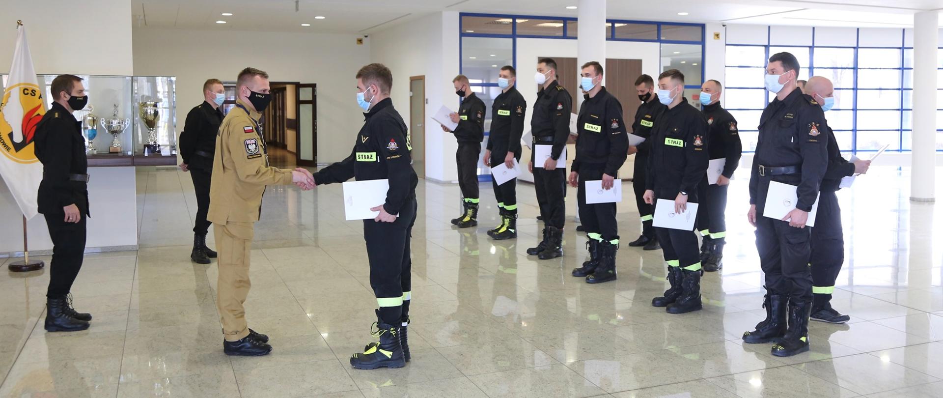 Zastępca Komendanta Centralnej Szkoły PSP gratuluje uczestnikowi szkolenia, w tle pozostali uczestnicy kursu stojący w dwóch rzędach