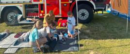 Na tle czerwonego wozu bojowego PSP grupka dzieci pod bacznym okiem funkcjonariusza Państwowej Straży Pożarnej ćwiczą na fantomie resuscytacje krążeniowo - oddechową