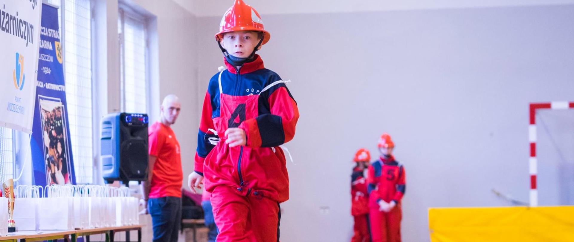 Na zdjęciu widać młodego uczestnika zawodów strażackich w hali sportowej. Dziecko ubrane jest w czerwony kombinezon z numerem „4” i pomarańczowy hełm ochronny, idzie po wąskiej drewnianej belce, będącej częścią toru przeszkód. W tle stoją inni uczestnicy w podobnych strojach, czekający na swoją kolej. Po lewej stronie widoczne są stoły z białymi torbami upominkowymi i pucharami oraz banery z logo powiatu wodzisławskiego i Ochotniczej Straży Pożarnej Bluszczów. Całość odbywa się w jasnej sali gimnastycznej z dużymi oknami.