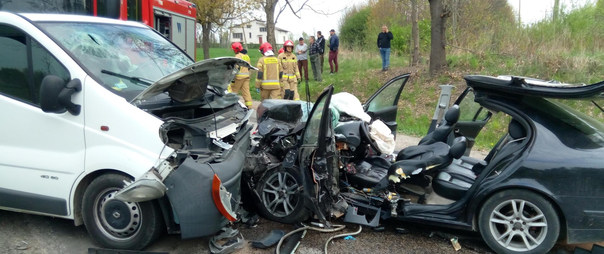 Na zdjęciu przedstawiono dwa auta po zderzeniu. Widać rozległe uszkodzenia obu pojazdów i odcięty dach w Seacie. Za samochodami znajdują się ratownicy a za nimi stoi grupa gapiów. Z lewej zaparkowany jest samochód pożarniczy.