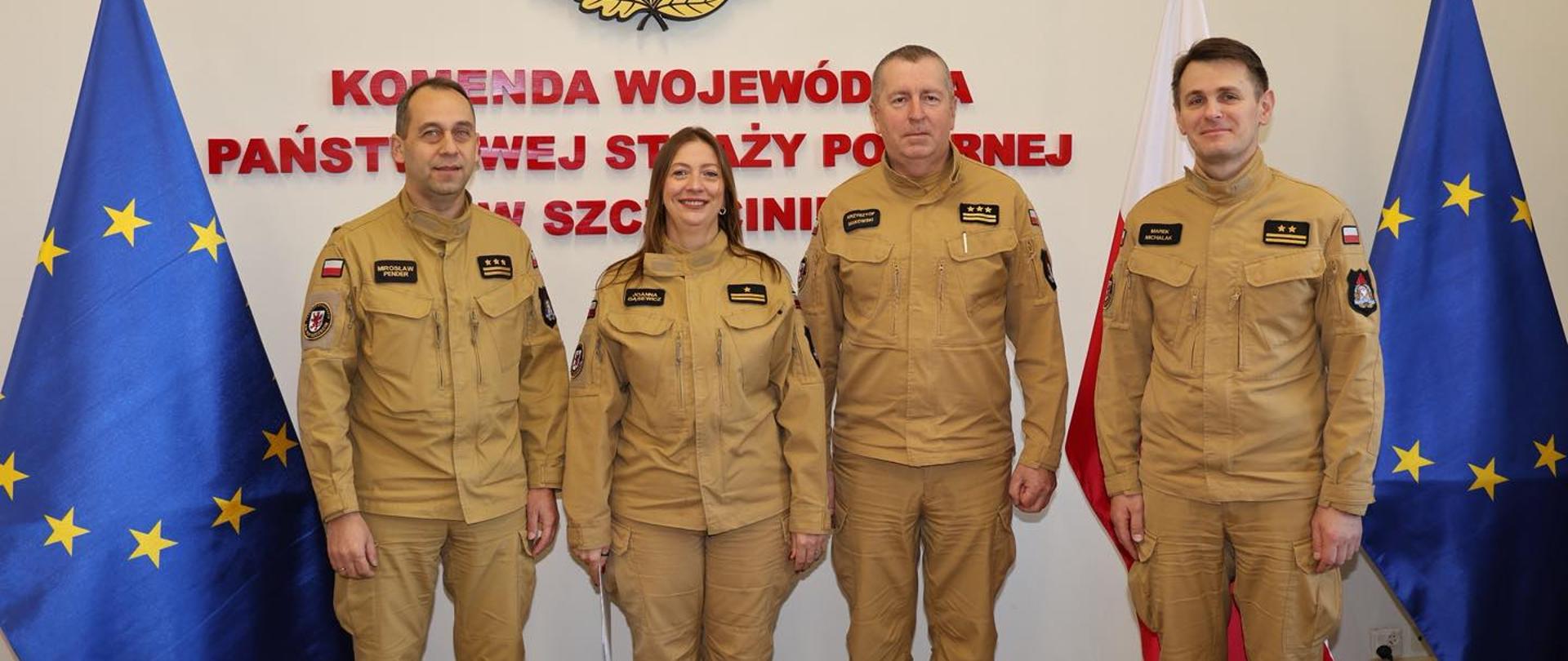 Na zdjęciu widać cztery osoby w piaskowych mundurach Państwowej Straży Pożarnej stojące na tle ściany z emblematem PSP oraz czerwonym napisem „Komenda Wojewódzka Państwowej Straży Pożarnej w Szczecinie”. Osoby stoją obok siebie, zwrócone twarzami do aparatu i uśmiechnięte. Po obu stronach grupy znajdują się stojące flagi: z lewej flaga Unii Europejskiej, z prawej flaga Polski oraz kolejna flaga Unii Europejskiej. Nad napisem widoczne jest godło PSP z orłem w czerwonym tle. Zdjęcie ma charakter oficjalny.