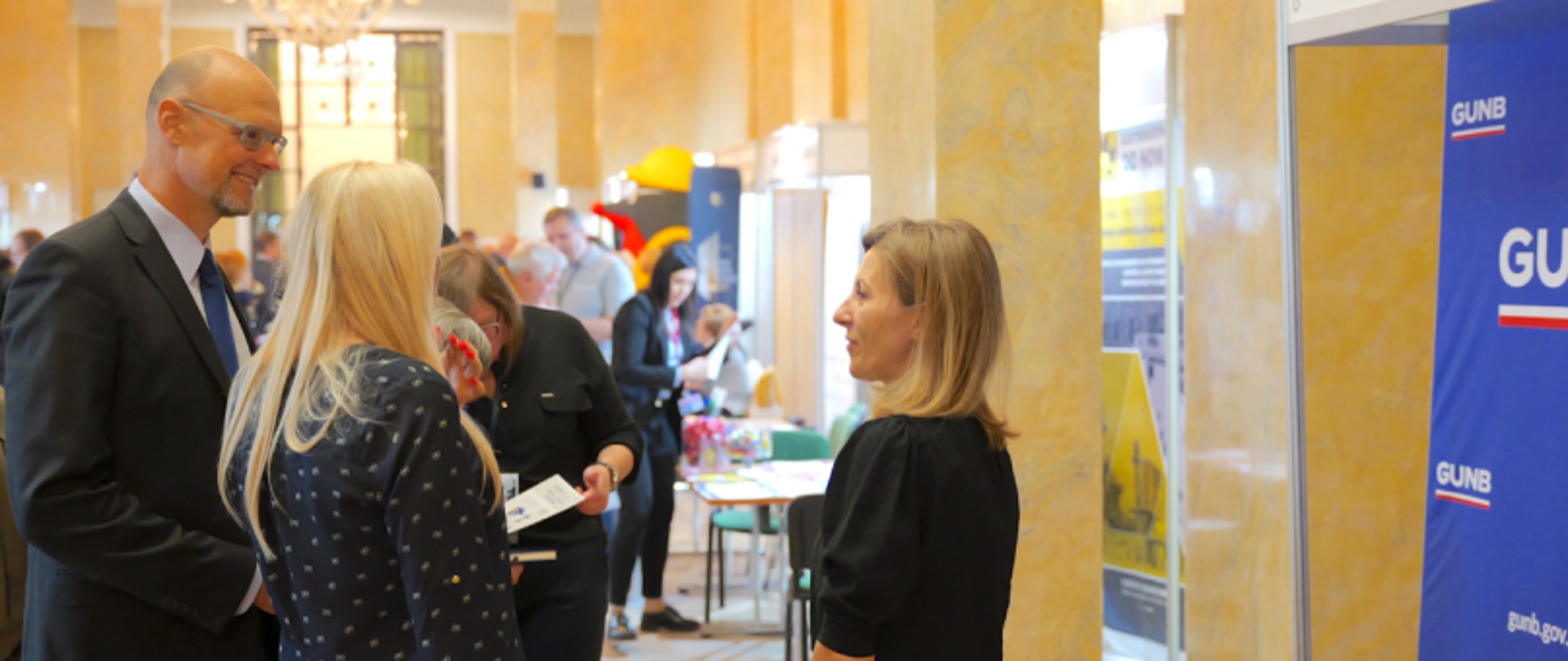 Stoisko Głównego Urzędu Nadzoru Budowlanego (GUNB) podczas targów pracy Talent Days. Przedstawiciele GUNB rozmawiają z uczestnikami wydarzenia na tle niebieskiej ścianki z logo urzędu. W tle widać innych odwiedzających i stoiska targowe.