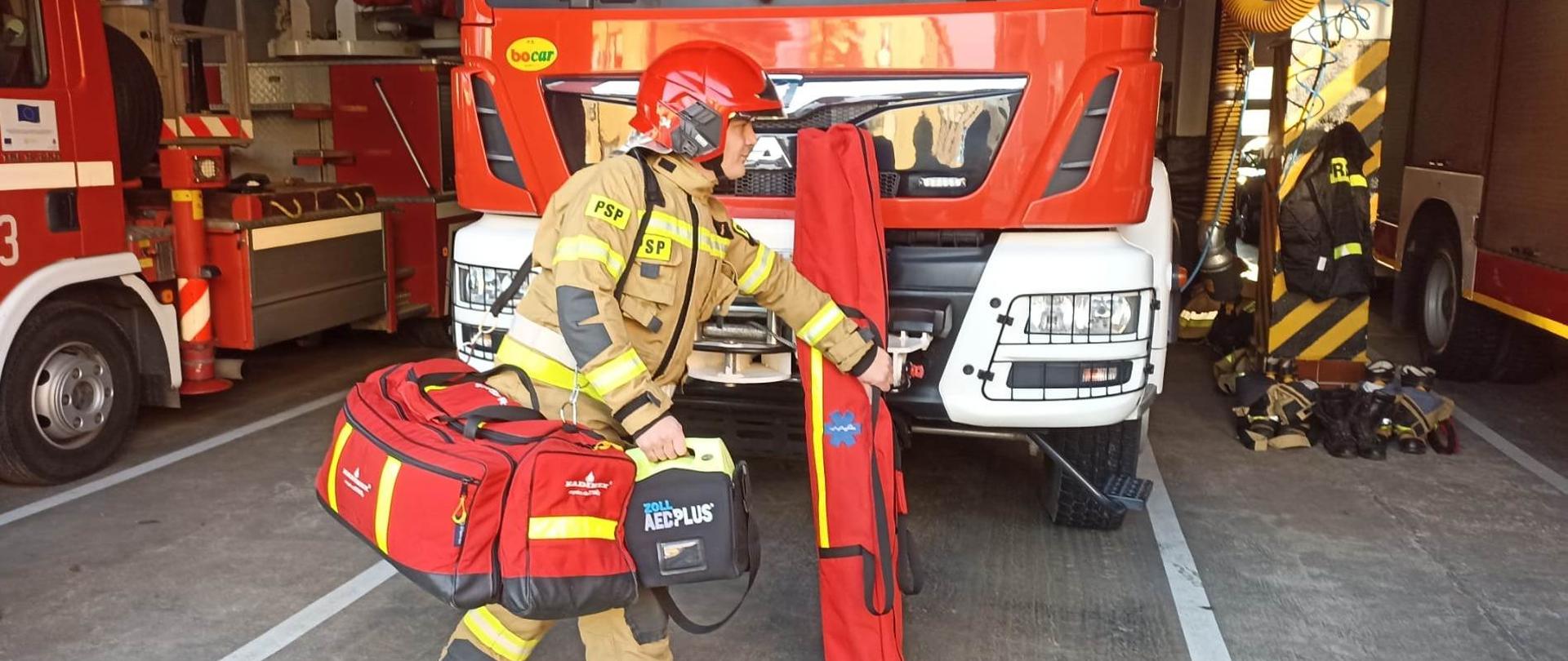 Kolorowa fotografia wykonana w dzień, w garażu straży pożarnej. Przedstawia na pierwszym planie strażaka w ubraniu specjalnym, w czerwonym hełmie na głowie, niosącego ratowniczy zestaw medyczny. Na drugim planie widoczna czerwona kabina stojącego pojazdu ratowniczo-gaśniczego znajdującego się w garażu. Obok widoczne częściowo inne pojazdy pożarnicze.