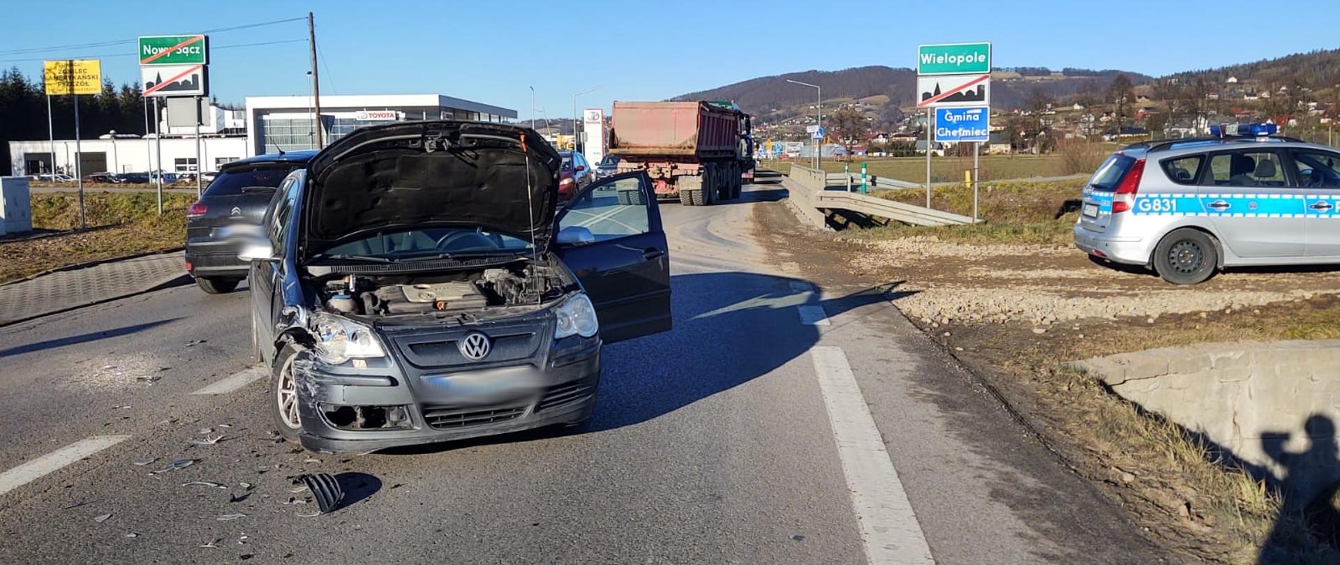 Samochód VW z otwartą klapą silnika stoi na środku drogi utrudniając przejazd. Na poboczu stoi radiowóz Policji.