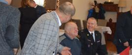 Świąteczne spotkanie strażackich emerytów. Sala w jednej z rawickich restauracji. Przy stole siedzi dwóch uczestników, w tym jeden w mundurze strażackim. Obok nich w kraciastej marynarce, pochylony jest kolejny mężczyzna. Panowie prowadzą rozmowę. 
