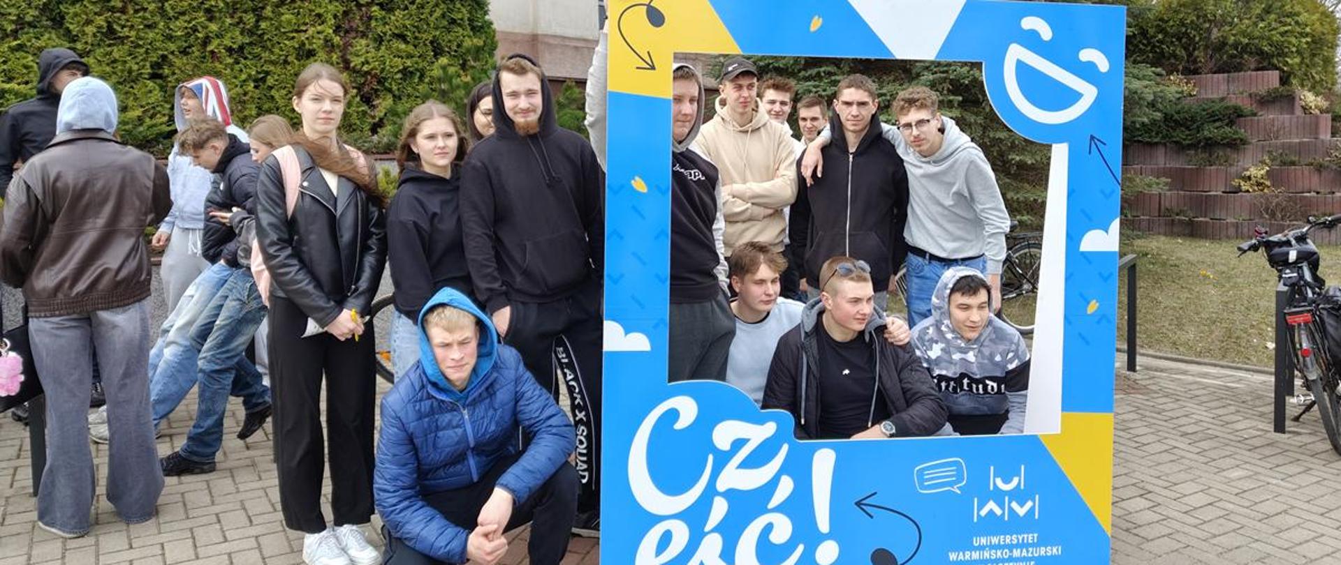 Grupa uczniów pozuje do wspólnego zdjęcia w dużej, ozdobnej ramie fotograficznej z napisem „UWM w Olsztynie” podczas wizyty na Dniu Otwartym uczelni.