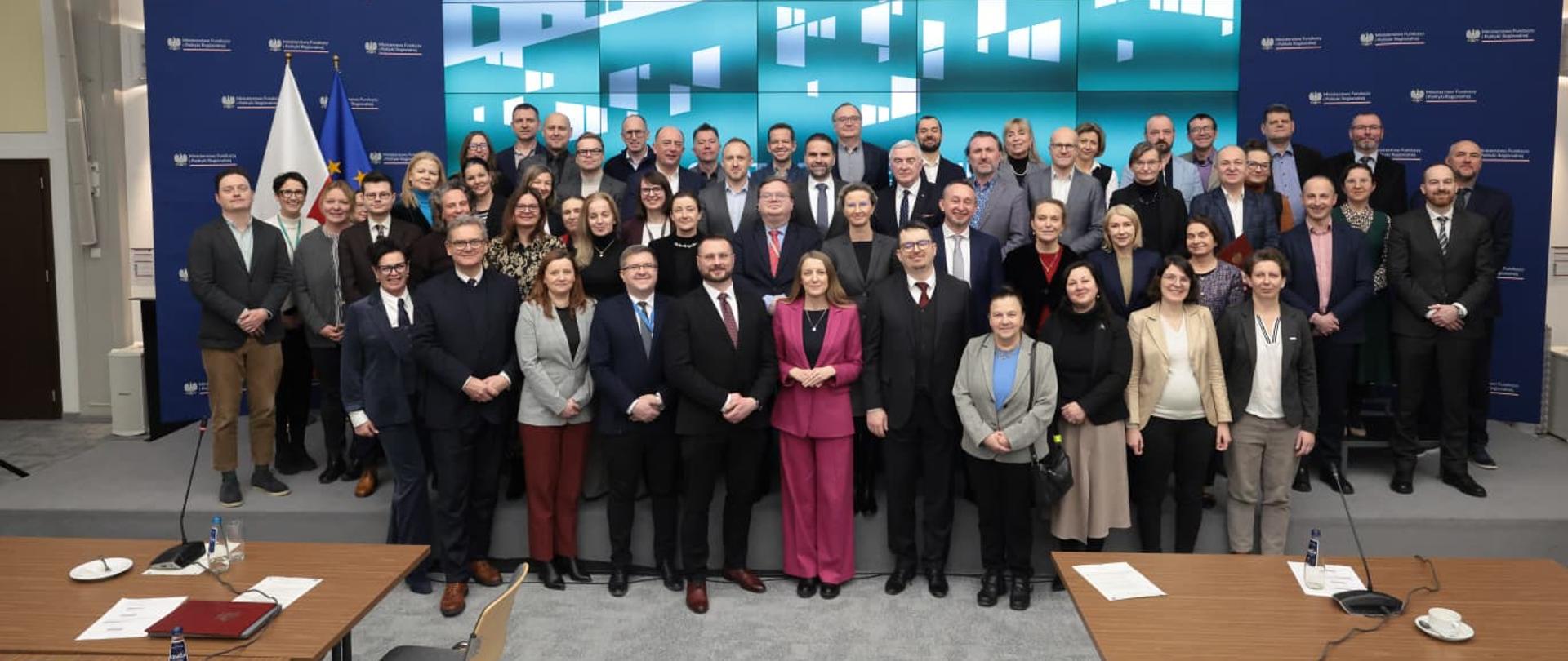 Na zdjęciu widać uczstników spotkania, duża grupa osób stoi na scenie w tle widać baner z grafiką projektu