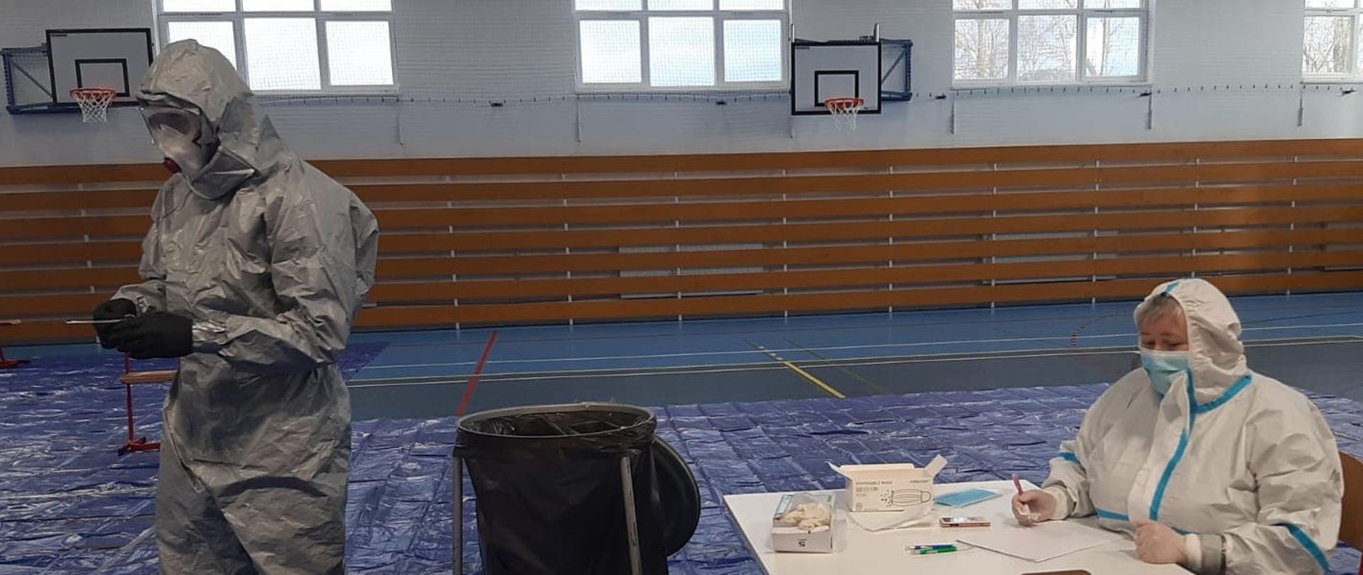 1. Fotografia została zrobiona na szkolnej sali gimnastycznej. W tle widać ścianę, na której zawieszone są dwie tablice do gry w koszykówkę oraz okna, poniżej nich znajdują się kaloryfery zabezpieczone drewnianymi deskami. Na podłodze została rozłożona niebieska folia. W lewej części fotografii widzimy strażaka ubranego w szczelny szary kostium, na jego twarzy znajdują się duże okulary ochronne oraz maska medyczna a czarne rękawiczki przykrywają dłonie,
w których trzyma pałeczki do wymazu. W centralnej części fotografii znajduję się duży kosz na odpady. Po prawej stronie można zobaczyć kobietę siedzącą za stołem, ona również ubrana jest w kombinezon ochronny oraz maseczkę i rękawiczki. Na stole znajdują się wymazówki, środki ochronne, dokumenty oraz taca z testami.