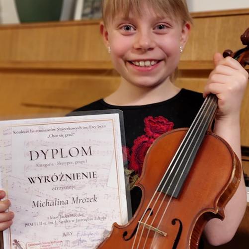 Uśmiechnięta dziewczynka trzyma w prawej ręce dyplom wyróżnienia, a w lewej skrzypce