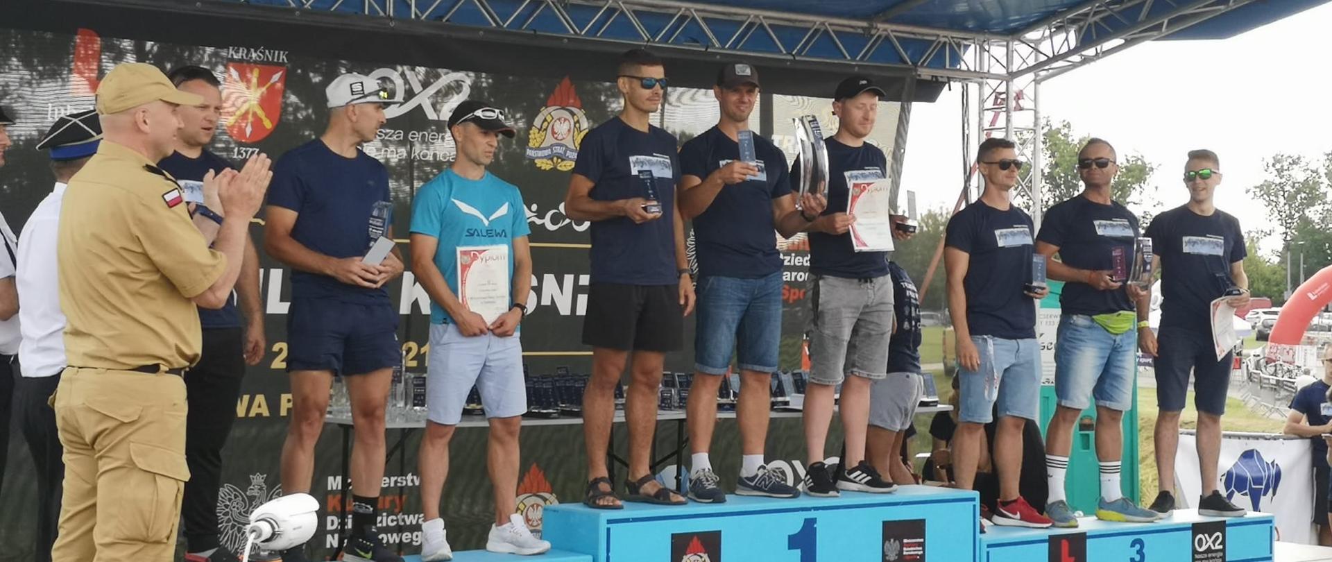 Zdjęcie przedstawia podium zawodów ustawione pod niebieskim zadaszeniem. Na każdym stopniu stoi 3 mężczyzn z trofeami. Z boku strażak w piaskowym mundurze bije brawo. 