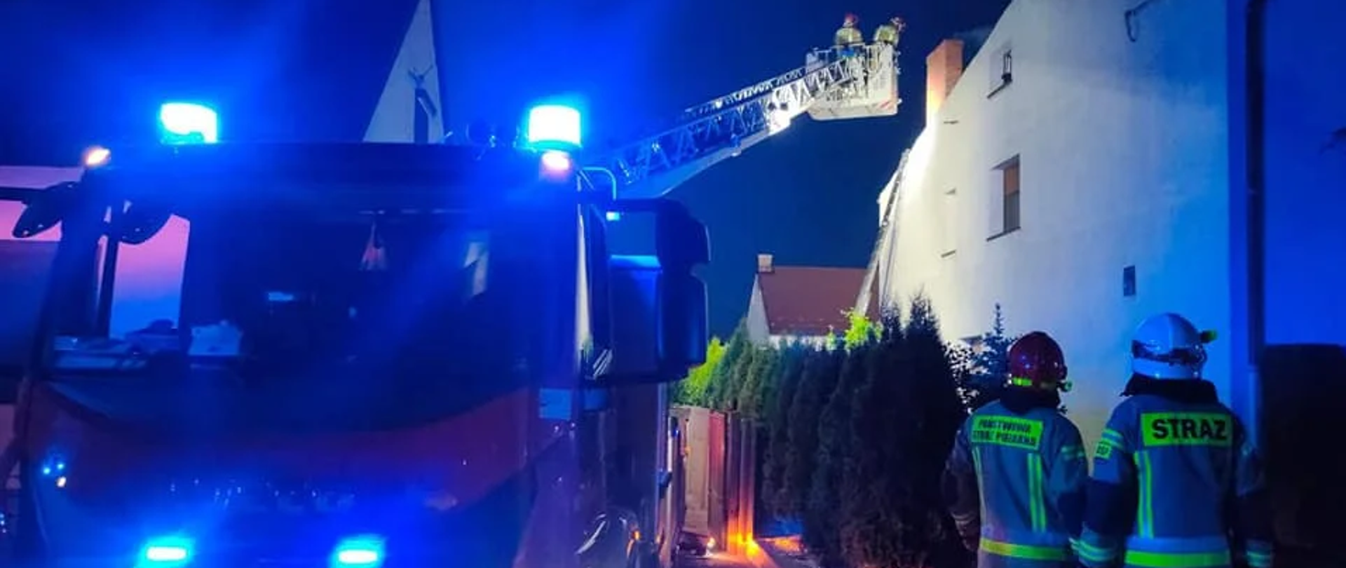 Zdjęcie przedstawia samochód pożarniczy z rozłożoną drabina. Przy samochodzie stoją strażacy.