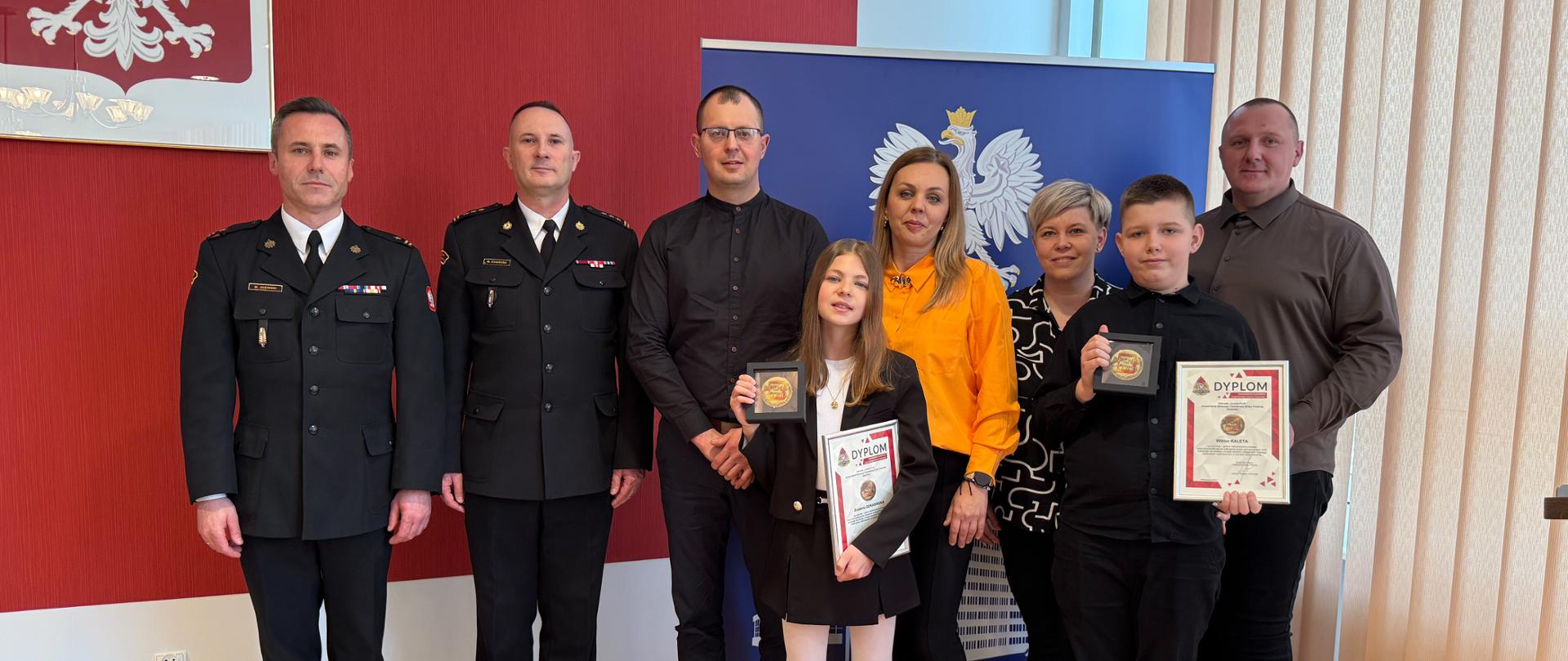 Na zdjęciu widoczny jest Świętokrzyski Komendant Wojewódzki Państowej straży pożarnej starszy brygadier Marcin Charuba oraz Komendant Powiatowy Państowej straży pożarnej w ostrowcu Świętokrzyski młodszy brygadier Marcin Jasiński wraz z młodymi bohaterami podczas odbioru podziekowań za ich bohaterski czyn.