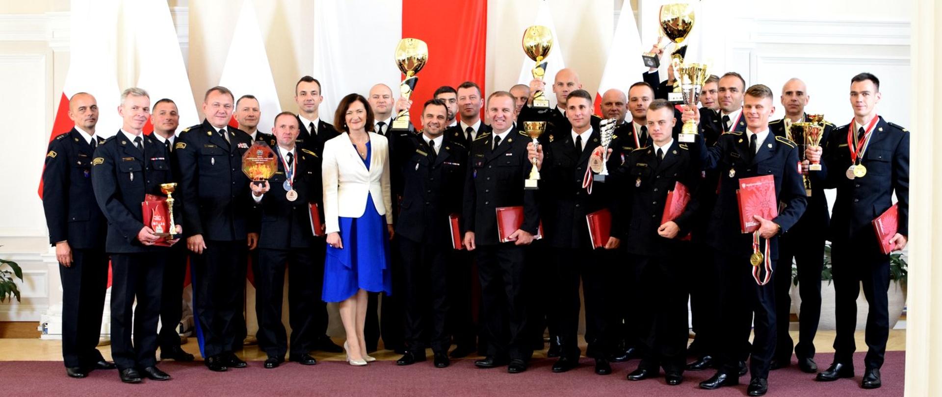Zdjęcie zrobione wewnątrz pomieszczenia, w sali kolumnowej podkarpackiego Urzędu Wojewódzkiego, podczas uroczystości wręczenia nagród strażakom. Na tle biało-czerwonych flag stoją wyróżnieni strażacy wraz z wojewodą podkarpackim oraz generałem Państwowej Straży Pożarnej. Strażacy trzymają w rękach puchary, dyplomy i medale.