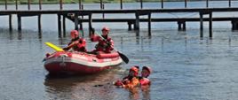 Grupa ratowników wodnych w czerwonych strojach i kaskach uczestniczy w ćwiczeniach na wodzie. W łodzi ratunkowej znajdują się dwie osoby, a jeden ratownik podejmuje pozoranta czekającego na pomoc w wodzie.