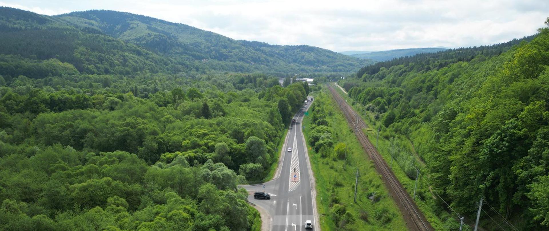 Widok z lotu ptaka na drogę krajową. Obok przebiega linia kolejowa, w otoczeniu zielone drzewa, a w tle góry