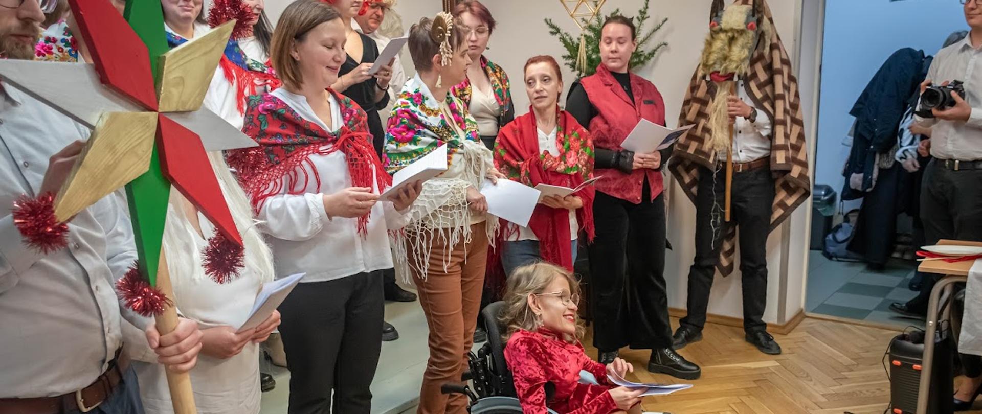 Zdjęcie przedstawia słuchaczy ubranych w kolorowe stroje i chusty, którzy w rękach trzymają kartki. Po środku jest słuchaczka na wózku inwalidzkim. 