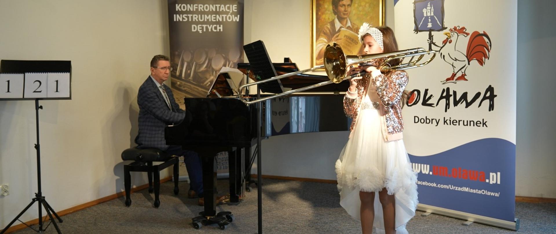 Uczennica gra na puzonie stojąc przed fortepianem, mężczyzna gra na fortepianie, w tle banery Konfrontacje Instrumentów Dętych oraz Urząd Miejski w Oławie - Oława dobry kierunek