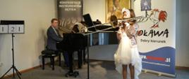 Uczennica gra na puzonie stojąc przed fortepianem, mężczyzna gra na fortepianie, w tle banery Konfrontacje Instrumentów Dętych oraz Urząd Miejski w Oławie - Oława dobry kierunek