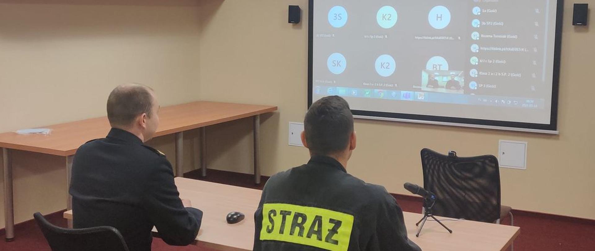 Przy biurku przed rzutnikiem siedzi dwóch strażaków jeden w mundurze granatowym drugi w czarnym. Z tyłu czarnego munduru napis STRAŻ 