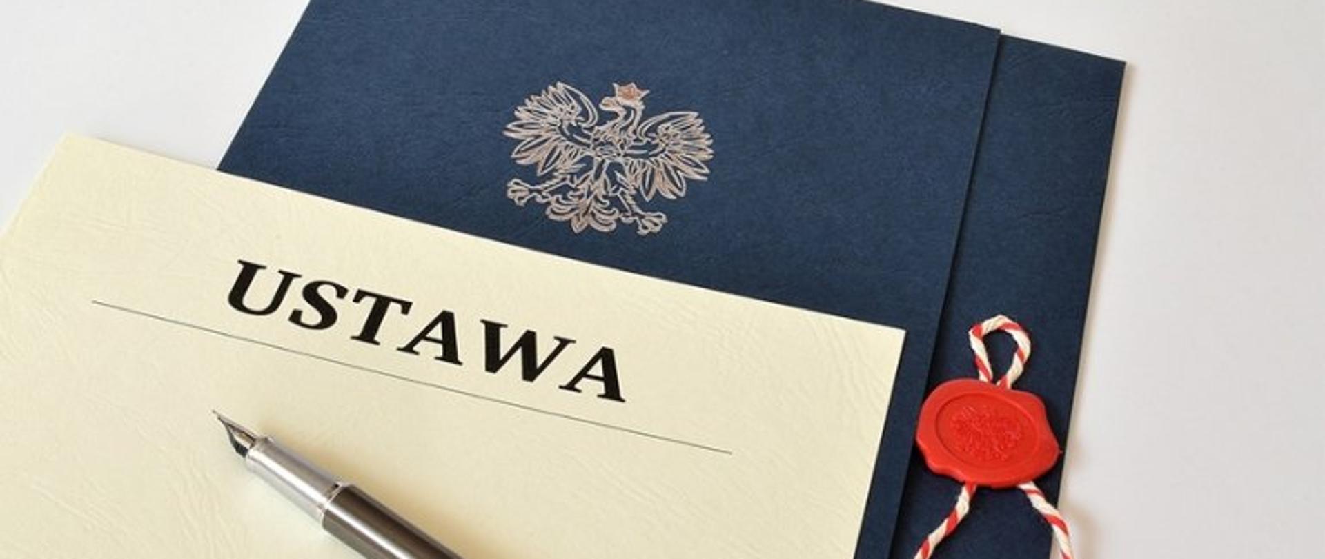 Na zdjęciu znajduje się granatowa teczka z białym nadrukiem godła i z pieczęcią. Na teczce leży biała kartka z napisem Ustawa oraz pióro.