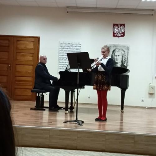 Zdjęcie przedstawia dziewczynkę grającą na trąbce, w tle akompaniator przy fortepianie. Na ścianie obrazy przedstawiające zapis nutowy oraz wizerunek patrona szkoły Jana Sebastiana Bacha.