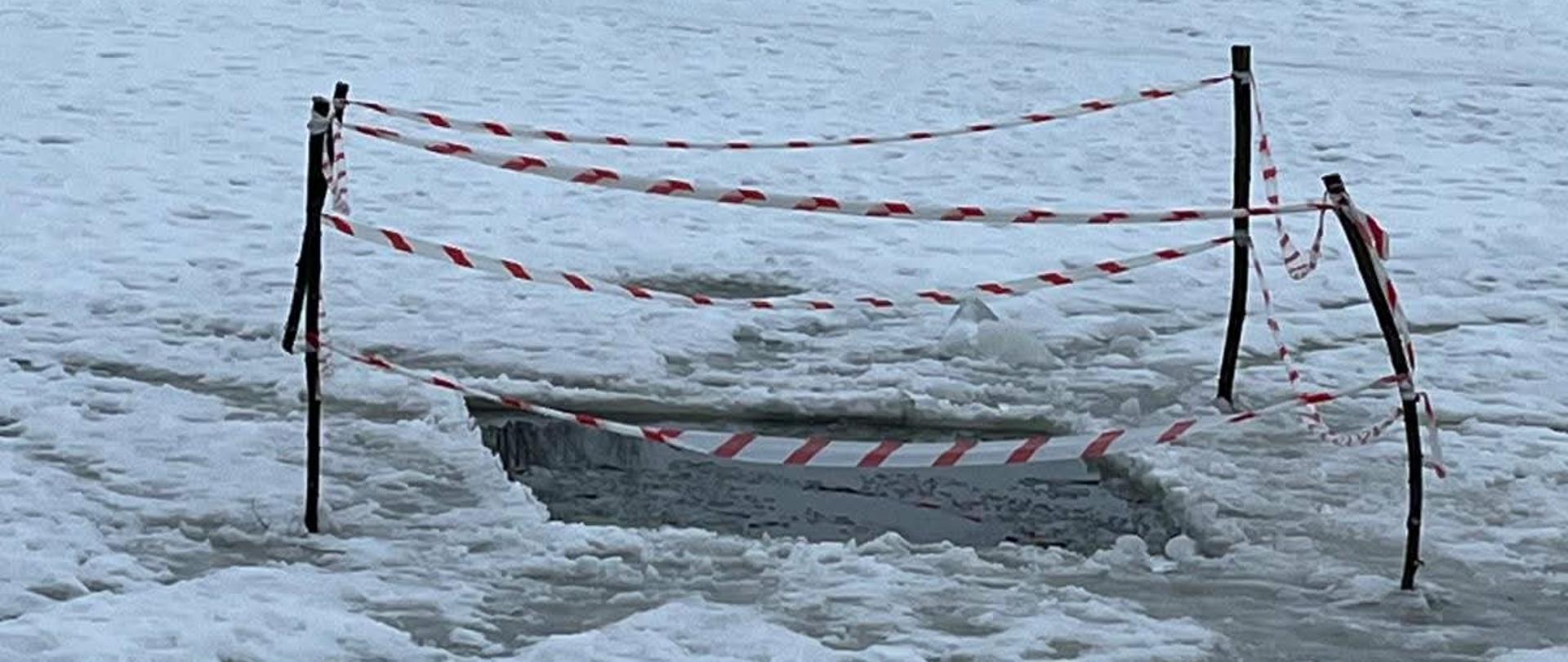 Na zdjęciu widać zimowy fragment parku, w którym część zamarzniętej powierzchni – prawdopodobnie stawu lub oczka wodnego – zaczęła topnieć. W miejscu, gdzie lód jest przerwany i widać wodę, ustawiono drewniane paliki z biało czerwoną taśmą ostrzegawczą, żeby nikt przypadkowo nie wszedł na niebezpieczny teren.
W tle stoją ławki, kosze na śmieci i niewielki biały budynek, a za nimi rosną drzewa. Całość sprawia wrażenie spokojnego, publicznego miejsca, w którym zadbano o to, by odwiedzający nie zbliżali się do osłabionego lodu.
