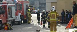 Ćwiczenia w ZOL. Zdjęcie przedstawia plac przed budynkiem zakładu opiekuńczo-leczniczego. Z lewej strony widoczne sa 2 wozy strażackie. Pomiędzy pojazdami a budynkiem znajdują sie strażacy biorący udział w ćwiczeniach. Maja na sobie ubrania specjalne w kolorze piaskowym i granatowym oraz białe, seledynowe i żółte hełmy. Przy pierwszym z samochodów ułożony jest sprzęt do ratownictwa medycznego. Do drugiego z samochodów podłączony jest biały wąż tłoczny, a do niego - rozdzielacz. 