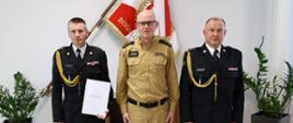 Trzech strażaków stoi obok siebie dwóch w mundurach wyjściowych ze sznurem w środku strażak w mundurze koloru musztardowego jeden z nich trzyma białą teczkę za nimi na ścianie wisi sztandar Komendy Głównej Państwowej Straży Pożarnej znajduje się szafka oraz po boku stoją donice z kwiatami.