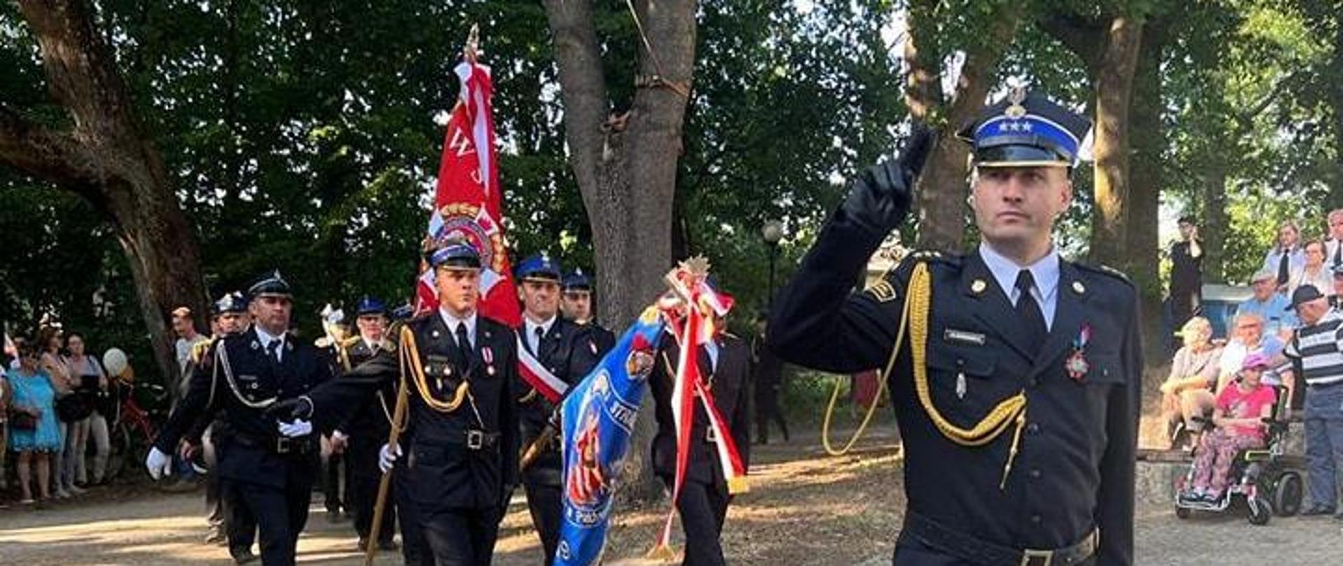 Zdjęcie przedstawia dowódcę pododdziałów - kpt. Grzegorza Sadowskiego podczas powiatowych obchodów Dnia Strażaka w Dobroniu. Za nim maszeruję poczty sztandarowe i poddziały biorące udział w uroczystości.