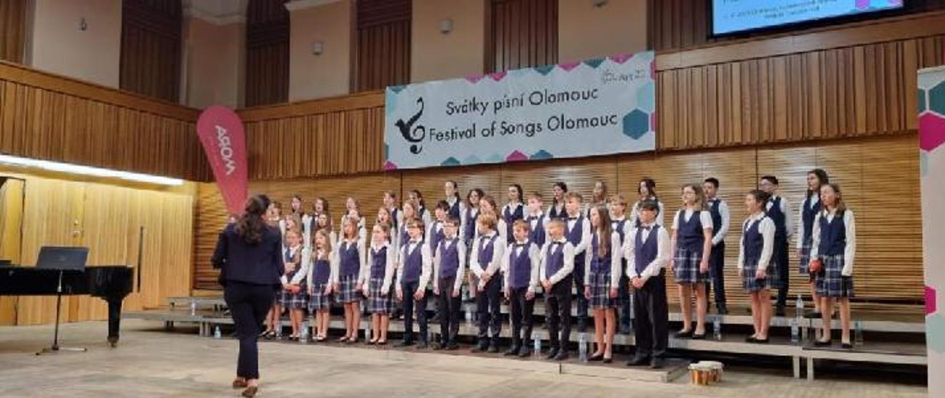 Na zdjęciu dzieci z chóru śpiewają na scenie, dyryguje pani Sobiecka.