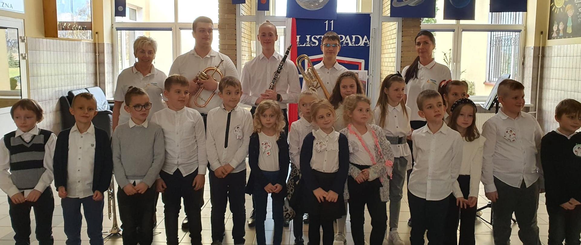 Uczniowie ubrani na galowo stoją przed tablicą z napisem 11 listopada, w drugim rzędzie nauczyciele i prowadzący z instrumentami: kornet, klarnet i puzon