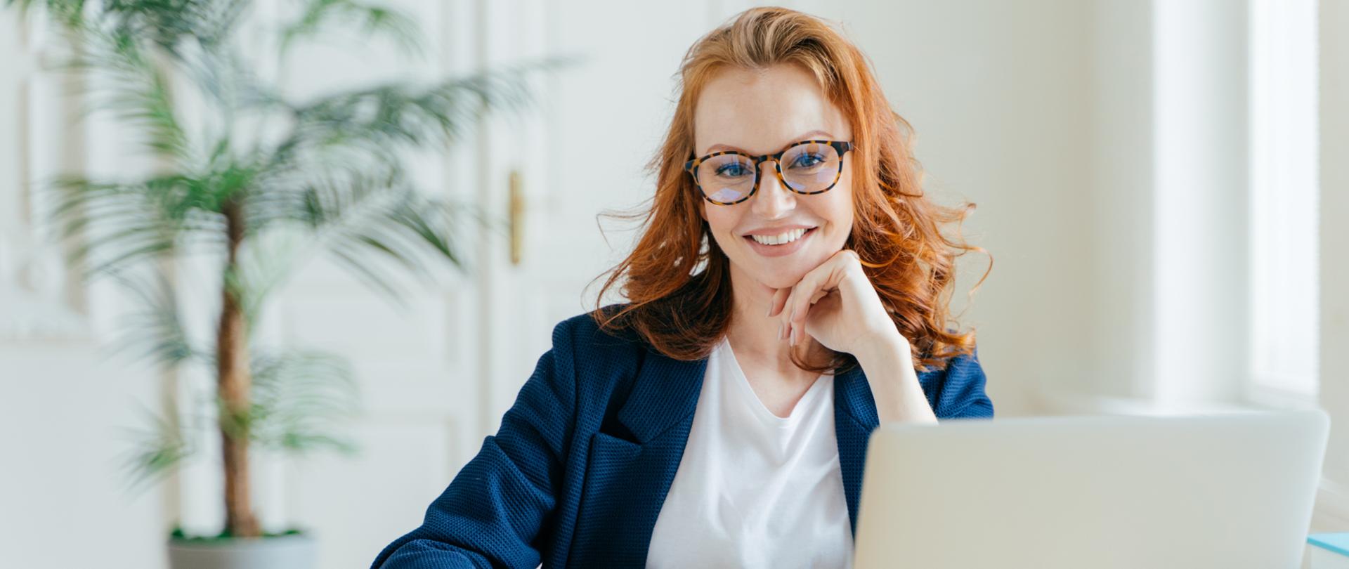 Zdjęcie rude kobiety, która siedzi przed laptopem i zapisuje informacje w notatniku