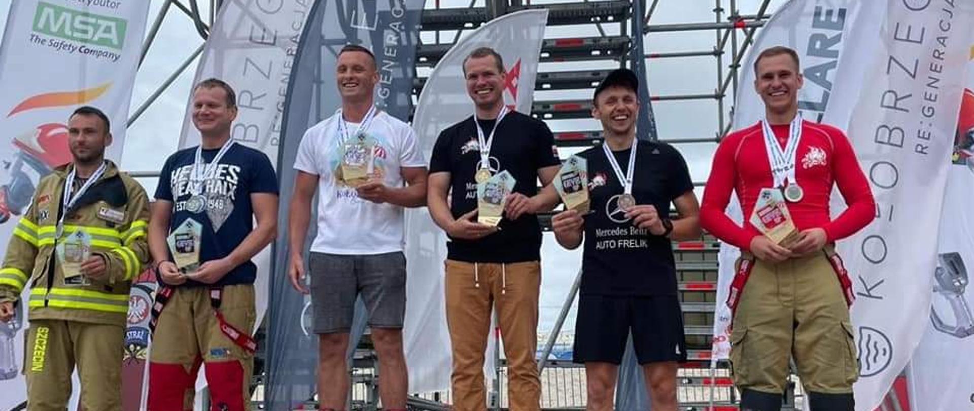 Podium, na którym stoją udekorowani strażacy. W tle flagi i bannery sponsorów z wieżą sportową. 