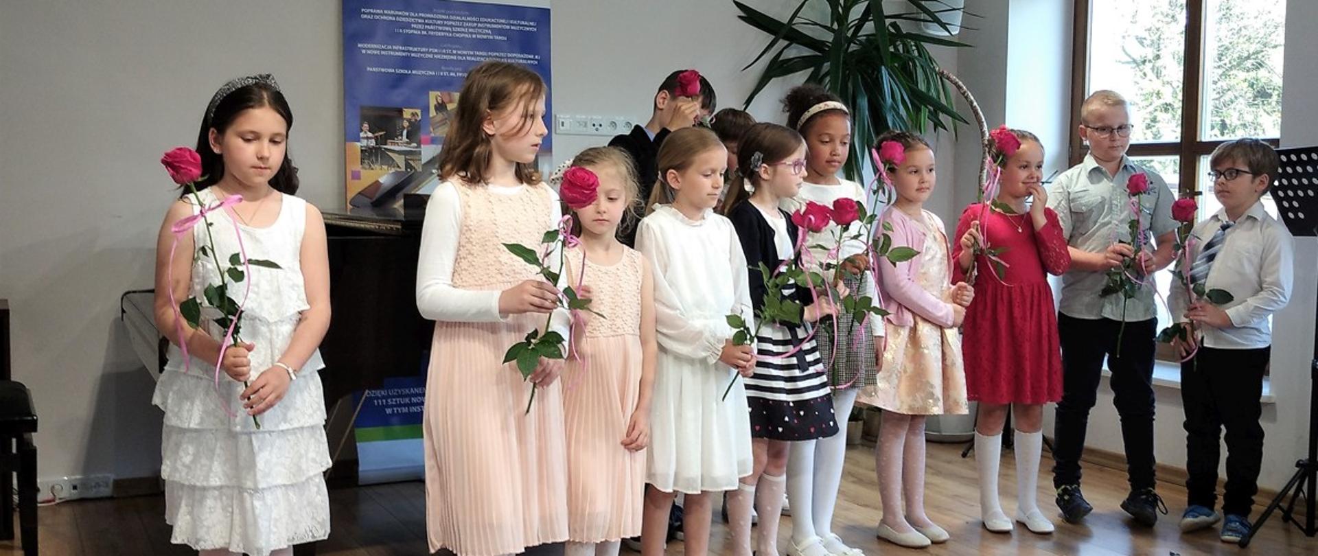 Na scenie sali koncertowej stoją najmłodsi uczniowie w rzędzie trzymają w dłoni róże. Za nimi fortepian, ława fortepianowa, baner unijny oraz palma doniczkowa. Po prawej stronie okno i jeden pulpit