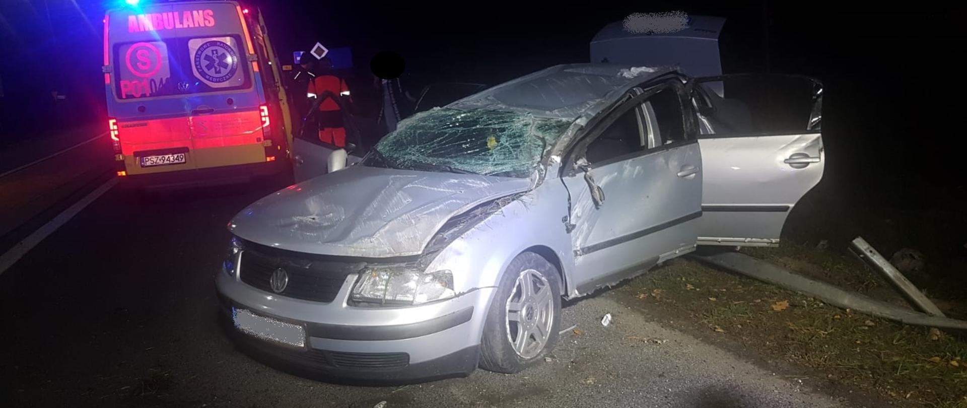 Widać uszkodzony samochód po dachowaniu, po lewej karetka pogotowia