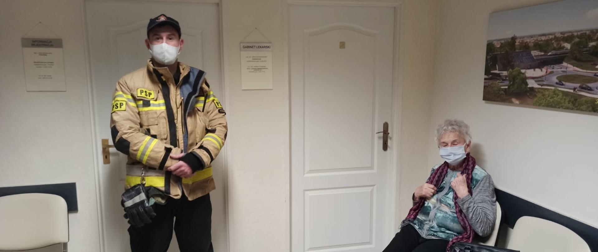Dwie osoby znajdujące się w korytarzu przychodni. W lewej części stojący strażak ubrany w żółtą kurtkę ubrania specjalnego i czarne spodnie, na twarzy biała maseczka. W prawej części starsza Pani siedząca na krześle ubrana jasny sweter i czarne spodnie, na twarzy maseczka. W tle ściany korytarza kremowego koloru i białe drzwi. 