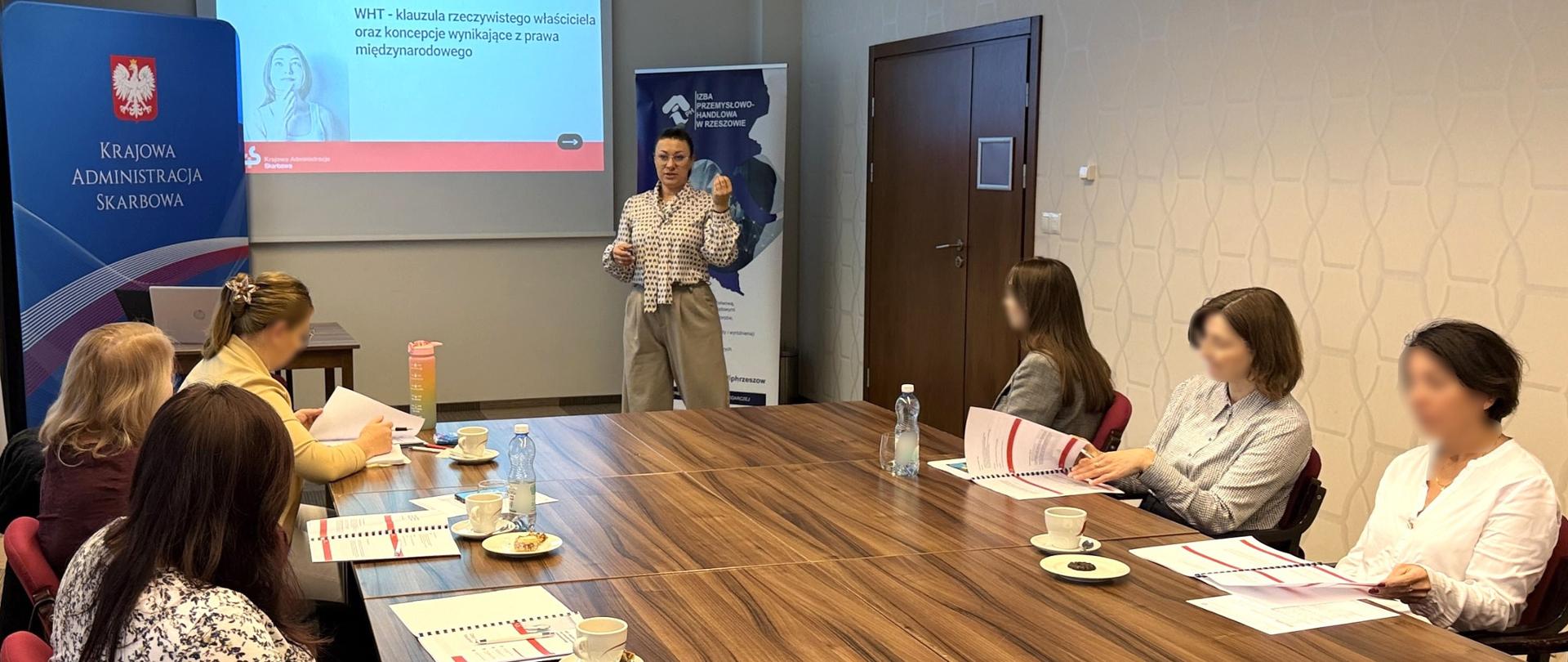 Uczestnicy szkolenia siedzą w sali. Przed nimi stoi ekspertka. Na ekranie wyświetlany jest slajd z napisem: WHT- klauzula rzeczywistego właściciela oraz koncepcje wynikające z prawa międzynarodowego.