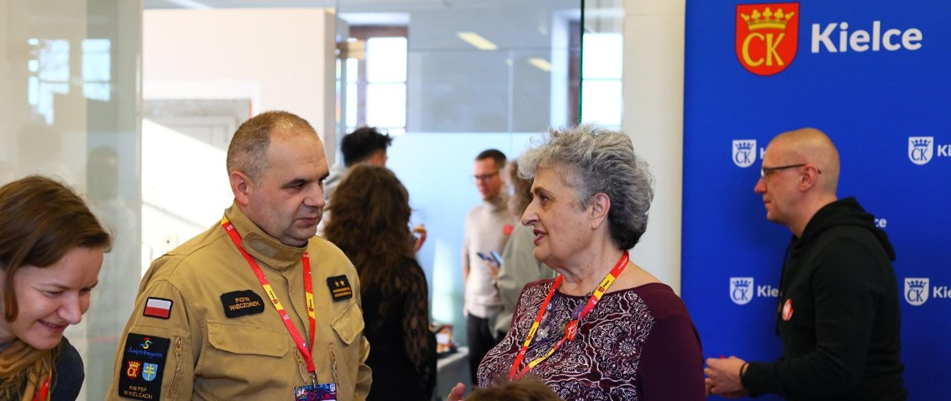 W pomieszczeniu podczas akcji charytatywnej „Malowanie serc” po lewej stronie stoi zastępca komendanta miejskiego PSP w mundurze, rozmawiający z kobietą z identyfikatorem. 