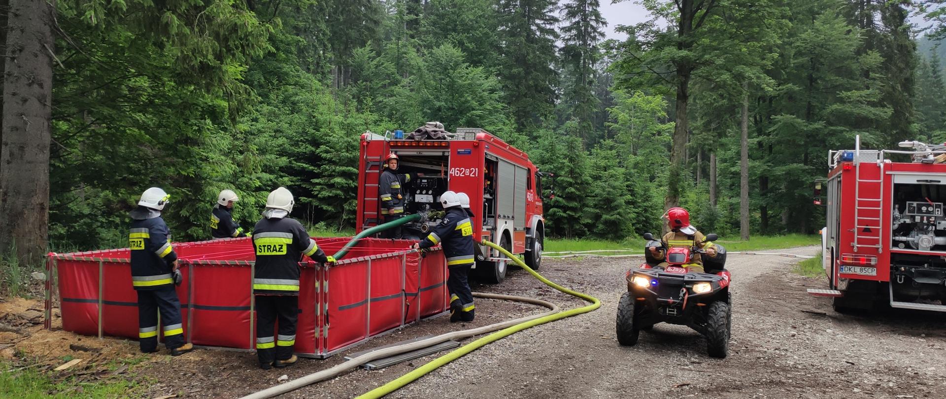 Strażacy w trakcie ćwiczeń. Na pierwszym planie droga leśna. Po lewej stronie strażacy pompują wodę ze zbiornika wodnego . Po prawej stronie siedzący na quadzie i obserwujący pompowanie wody strażak. W tle las..