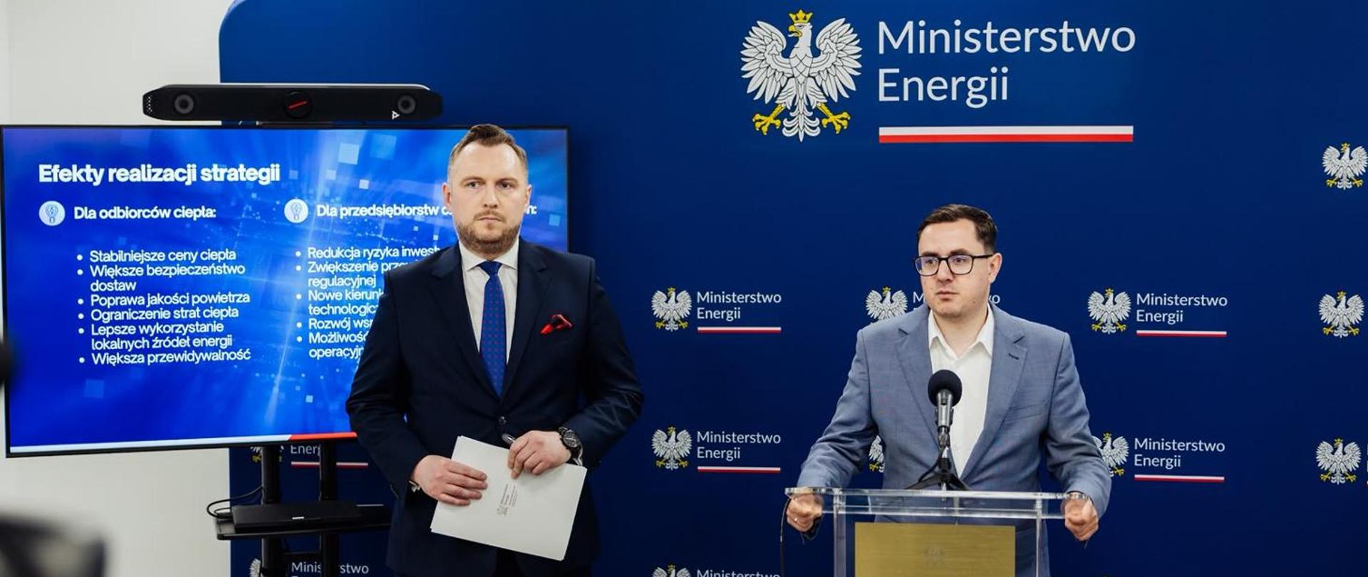 Ministrowie podczas konferencji Ministerstwa Energii stoi przy mównicy i ekranie z prezentacją o efektach strategii.
