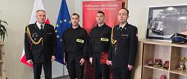 Czterech strażaków stoi w pomieszczeniu podczas uroczystości ślubowania. Dwóch w galowych mundurach i dwóch w umundurowaniu służbowym. W tle widoczne są flagi Polski i Unii Europejskiej oraz baner Państwowej Straży Pożarnej. Po prawej stronie znajduje się regał z pamiątkami i odznaczeniami.