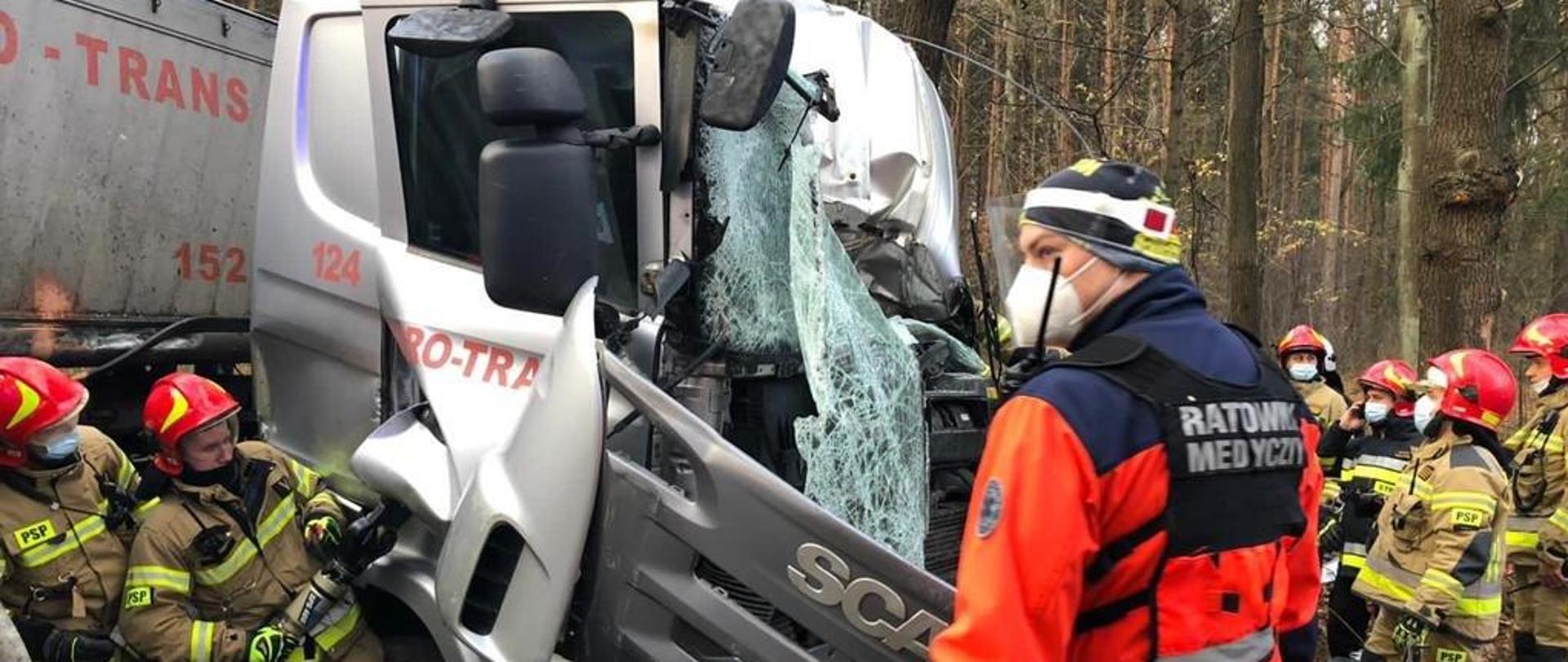 Zdjęcie przedstawia akcję ratowniczą podczas wypadku drogowego.
Po lewej stronie zdjęcia (na pierwszym planie) widoczny kawałek żółtego węża hydraulicznego. Na dalszym planie widoczni dwaj strażacy PSP (ubranie specjalne koloru piaskowego z czerwonym hełmem z maseczkami), którzy przy pomocy narzędzia hydraulicznego wyważają drzwi od strony pasażera w ciągniku siodłowym marki Scania koloru srebrnego. Za strażakami widoczny kawałek ustawionej w poprzek naczepy koloru szarego. Kabina ciągnika siodłowego z wybitą (stłuczona) przednia szyba i wgniecionym dachem od strony rogu kierowcy pojazdu. Na pierwszym planie, przed pojazdem widoczny ratownik medyczny ustawiony bokiem (ubranie jaskrawo pomarańczowe z kamizelką z napisem "ratownik medyczny") z półmaską, rękawiczkami nitrylowymi na rękach koloru niebieskiego oraz przyłbicy przezroczystej zasłaniającej całą twarz.
Po prawej stronie czterech strażaków PSP (trzeb w ubraniach specjalnych koloru piaskowego a jeden czarnego, wszyscy w czerwonych hełmach) stojący przodem do rozbitej kabiny ciężarówki. W tle zdjęcia las.