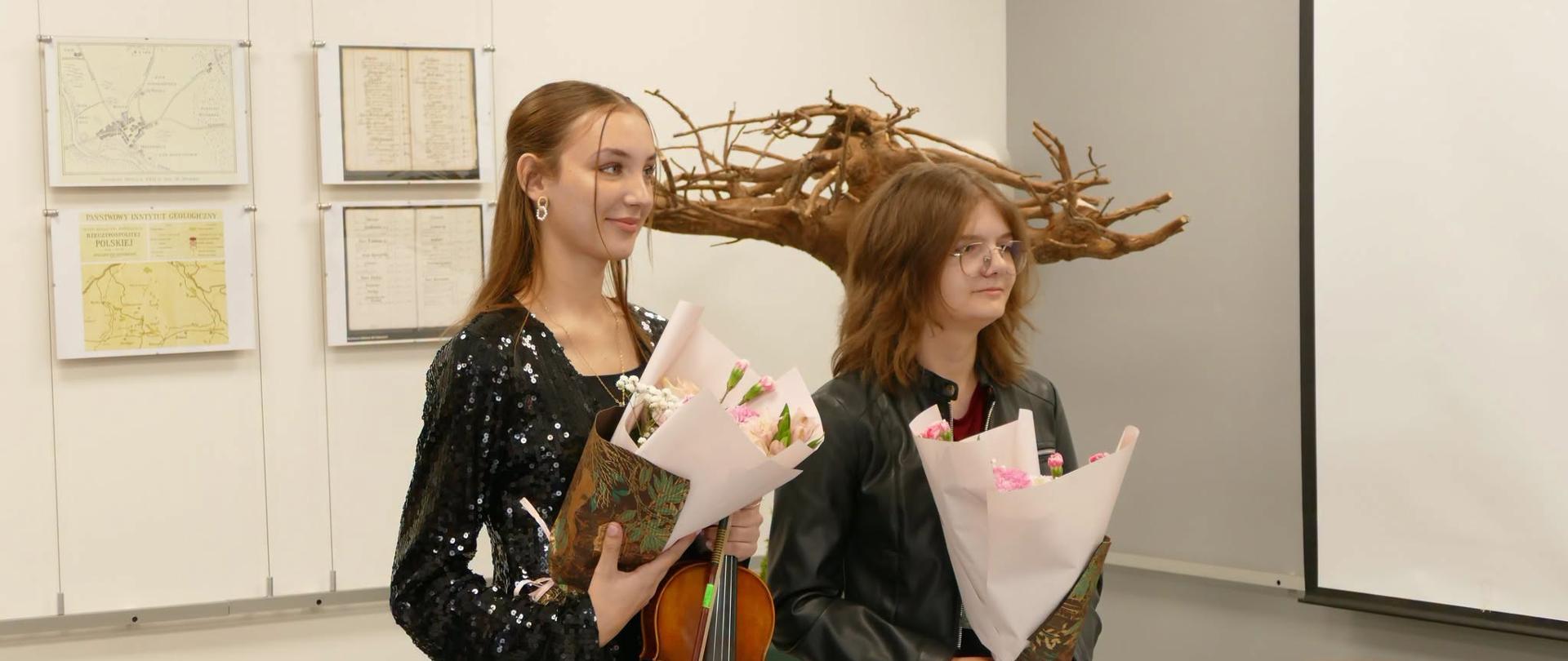 Julia Kurnal ze skrzypcami i bukietem kwiatów oraz Daria Kopacz z bukietem kwiatów stoją po występie w sali wystawowej Nadleśnictwa Mielec, w tle mapy i rzeźba z drewna.