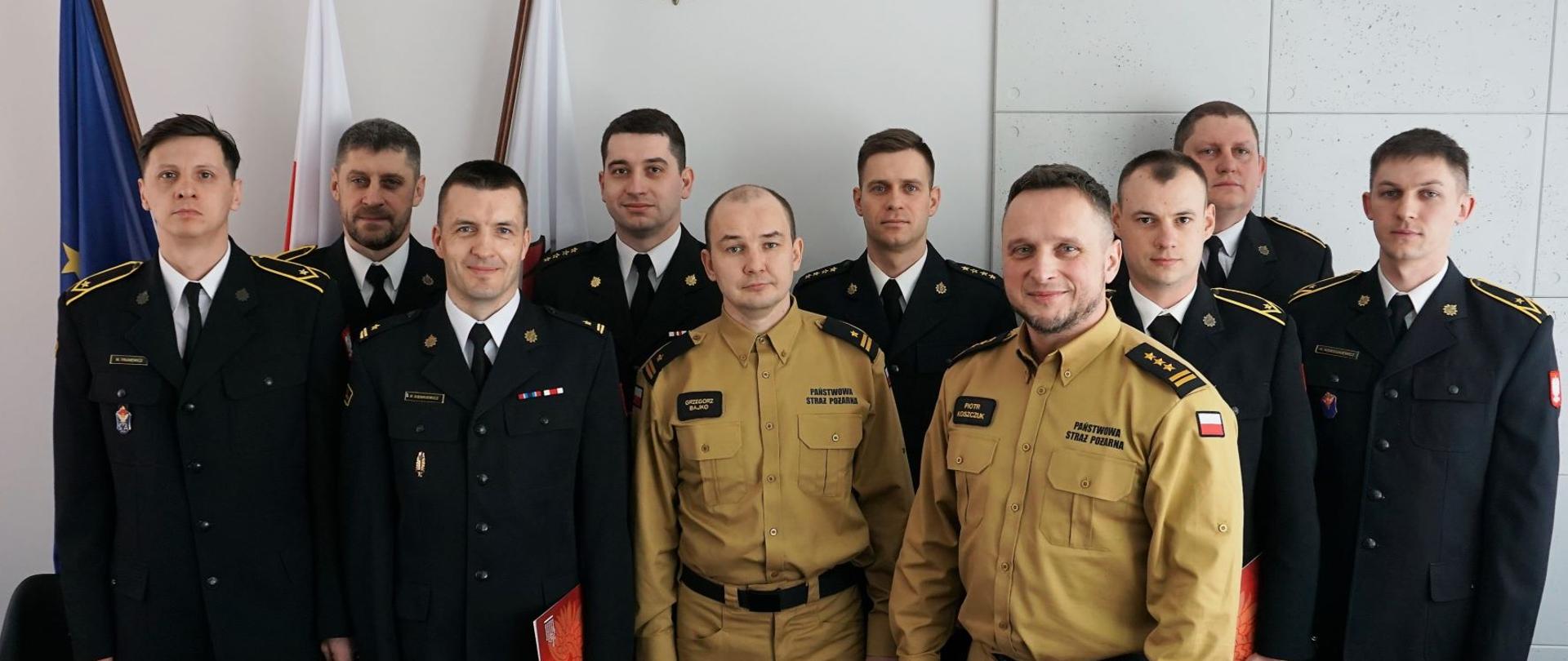 W pomieszczeniu w dwóch rzędach stoi dziesięciu strażaków w mundurach. U góry godło, obok flaga państwowa.