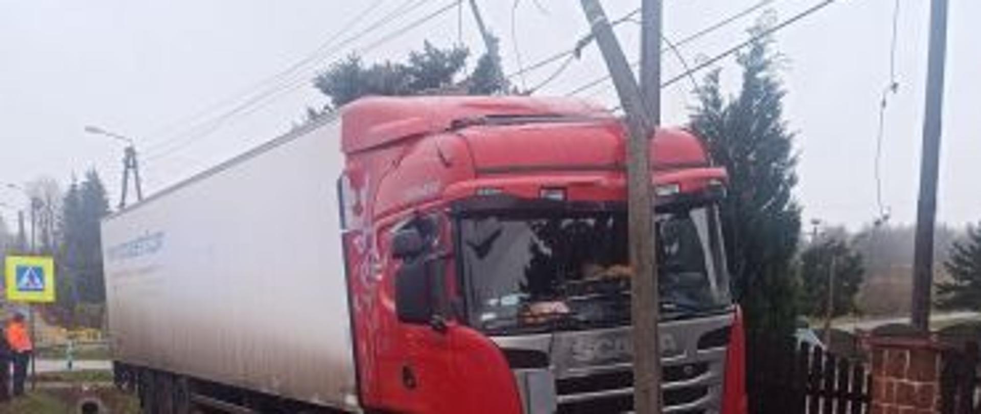 Samochód ciężarowy z naczepą zjechał z pasa drogi na chodnik i zatrzymał się przy ogrodzeniu prywatnej posesji. Pojazd centralnie uderzył kabiną w stojący słup energetyczny. Górna część słupa, która została złamana zawisła na przewodach nad kabiną kierowcy pojazdu. Za ogrodzeniem widać drzewa.