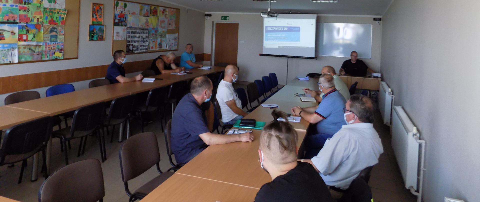 Zdjęcie przedstawia Komendanta Powiatowego Państwowej Straży Pożarnej w Rypinie starszego brygadiera Grzegorza Łydkowskiego, który podczas spotkania z druhami Ochotniczych Straży Pożarnych w świetlicy edukacyjnej Komendy Powiatowej, wyjaśnia założenia programu Szczepimy się z OSP.
Na zdjęciu widzimy łącznie dziesięć osób. W tle widnieje prezentacja programu Szczepimy się z OSP.