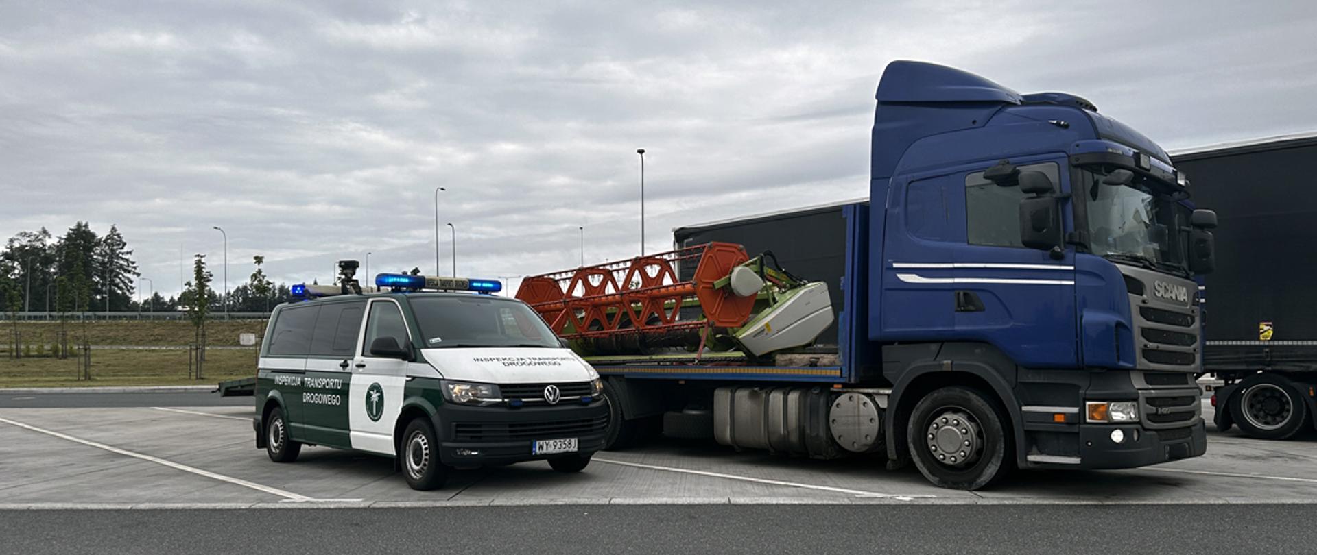 Na pierwszym planie znajduje się samochód ITD z włączonymi niebieskimi sygnałami świetlnymi, zaparkowany obok niebieskiej ciężarówki z naczepą. Na naczepie przewożony jest element maszyny rolniczej