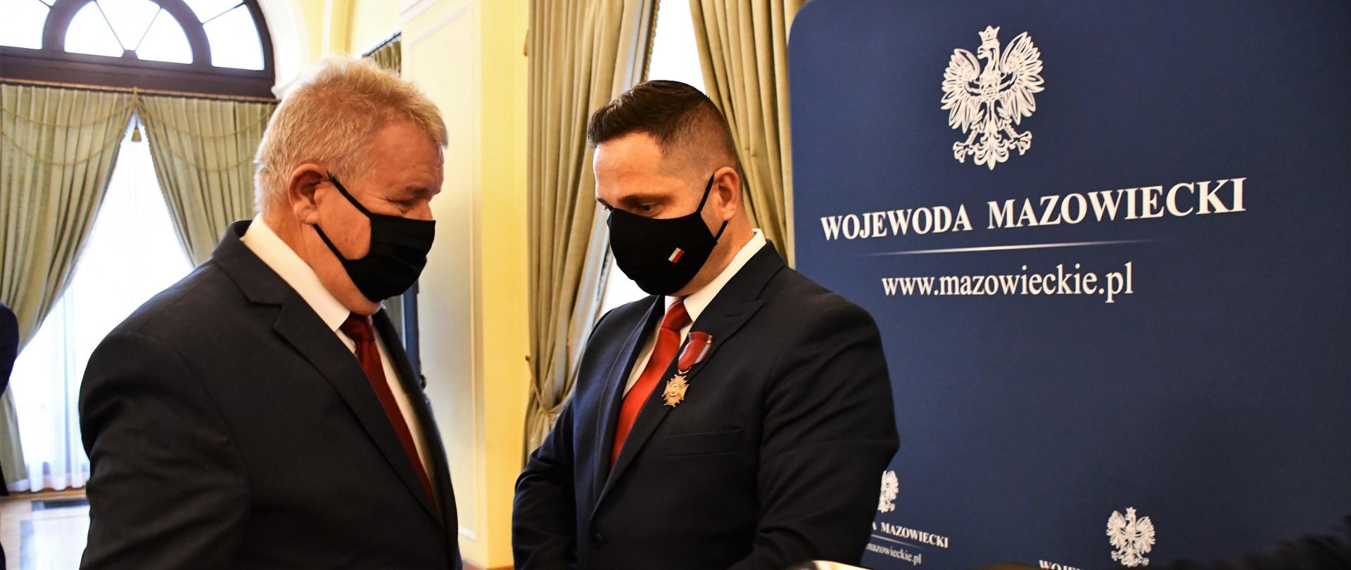 Fotografia przedstawia dwóch mężczyzn, ubranych w garnitury, białe koszule i krawaty. Jeden z mężczyzn odznaczony został Brązowym Krzyżem Zasługi, który przypięty jest w klapie marynarki. na drugim planie fragment sali konferencyjnej z wysokimi oknami oraz ścianka wizerunkowa z Orłem RP oraz napisem "Wojewoda Mazowiecki". 