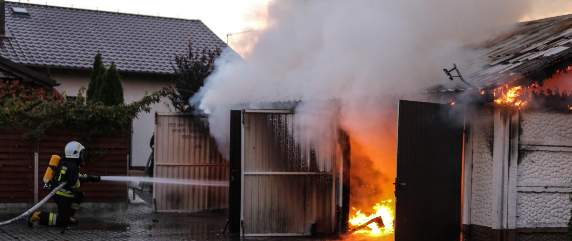 Zdjęcie przedstawia strażaka, który gasi pożar budynku garażowego dwustanowiskowego. Budynek wykonany z płyt betonowych, dach dwuspadowy wykonany z konstrukcji drewnianej pokryty blachą trapezową. Budynek cały w ogniu